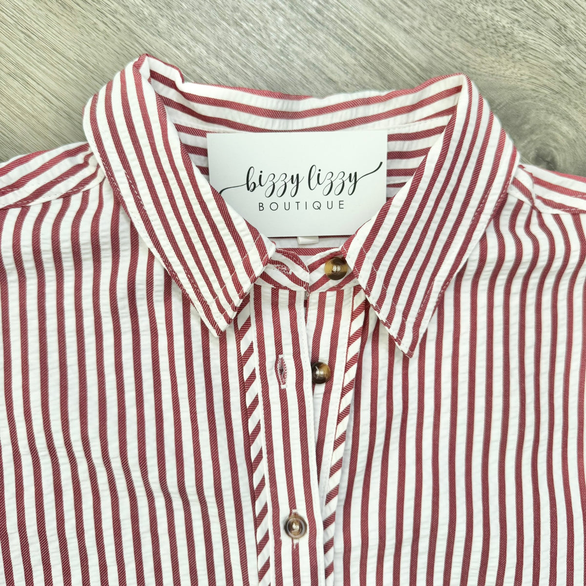 A close-up of a collared button-up shirt with vertical red and white stripes, featuring a white neck label that reads 'bizzy lizzy BOUTIQUE' and brown buttons, laid flat on a light gray wood-textured surface.