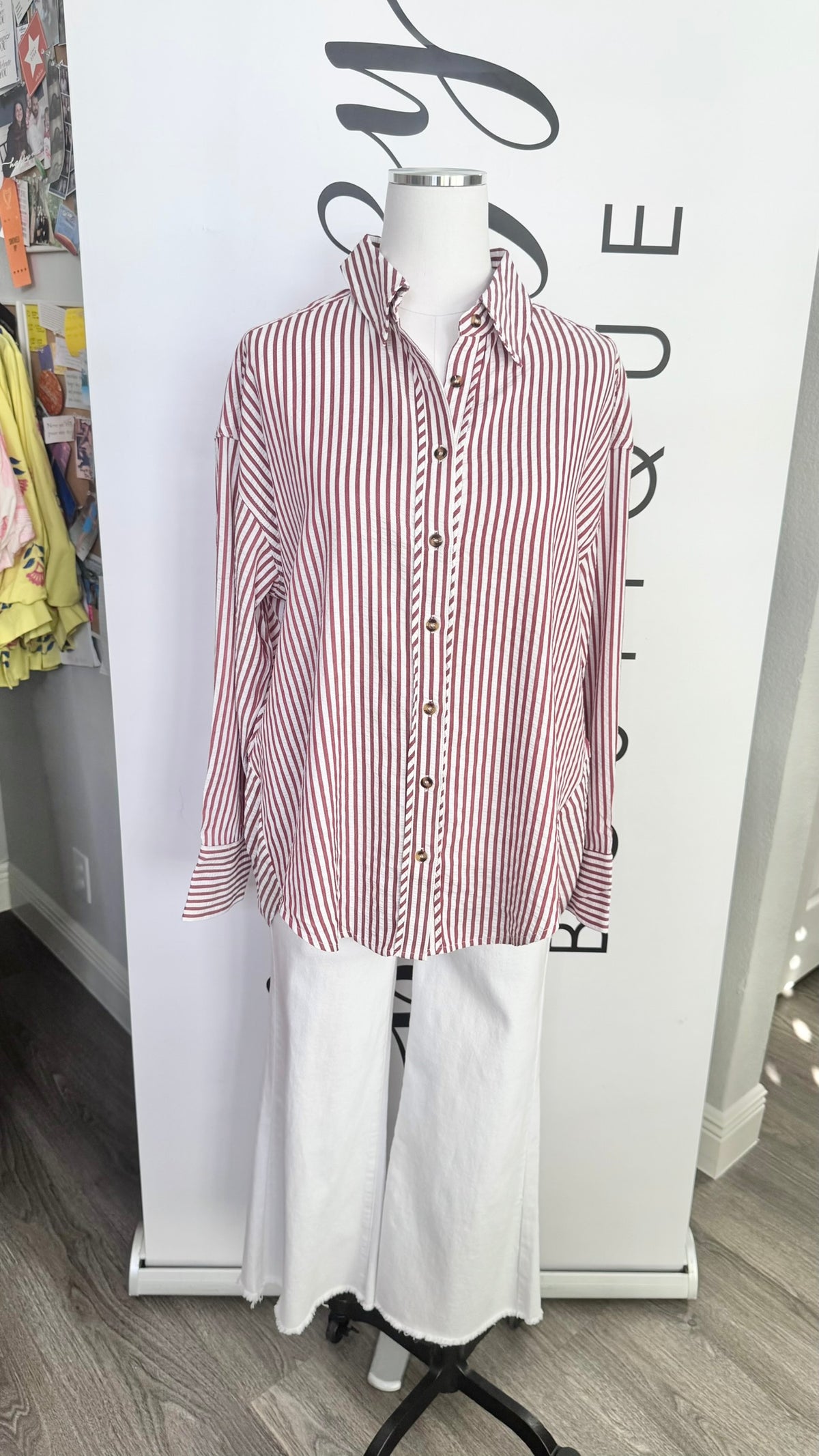 A long-sleeve button-up shirt with vertical red and white stripes, displayed on a white mannequin torso with white wide-leg pants. The mannequin is positioned in front of a white boutique display banner.