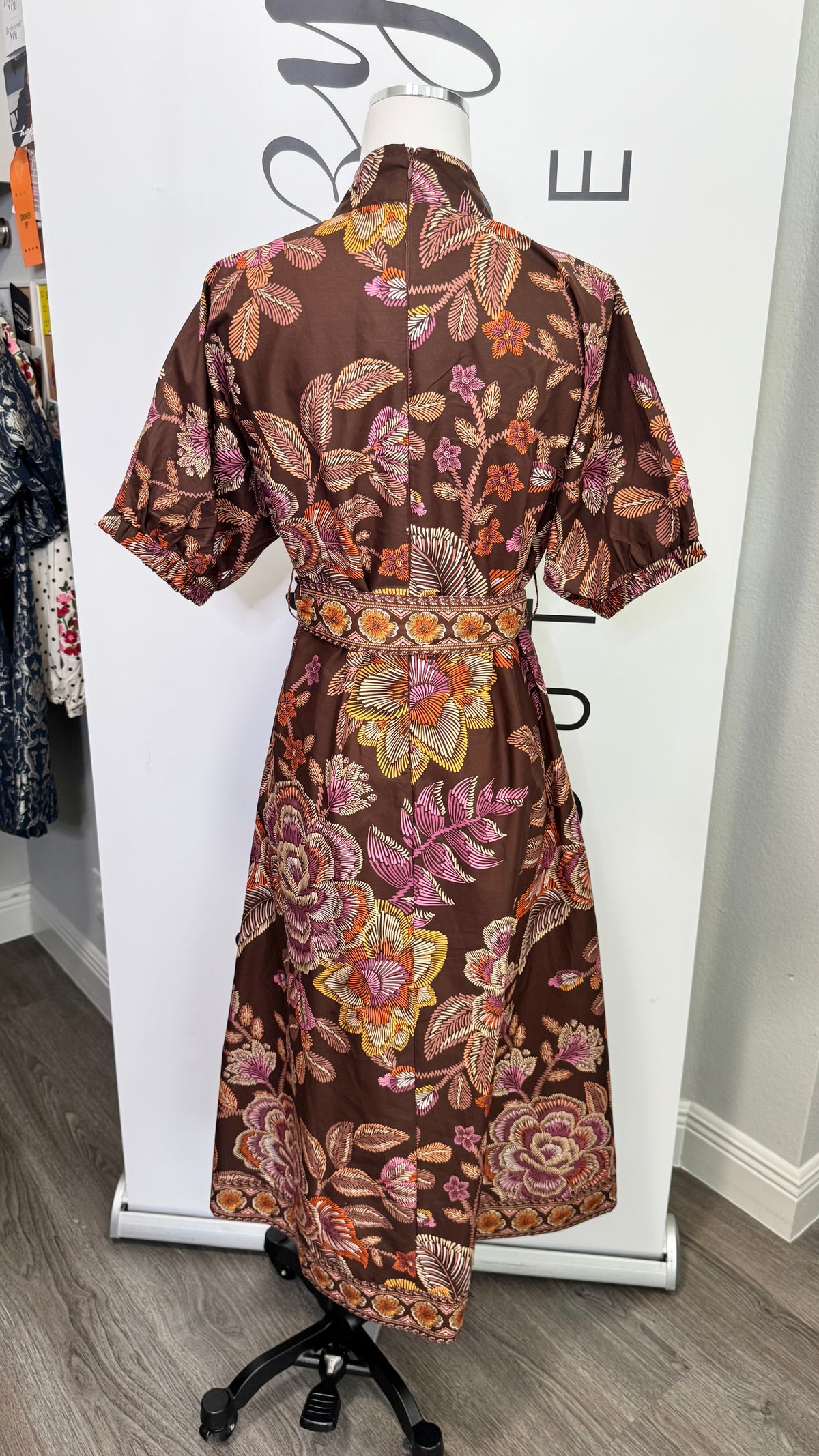 A brown short-sleeve belted dress with a vibrant pink, orange, and yellow floral print, displayed on a mannequin in a boutique setting.