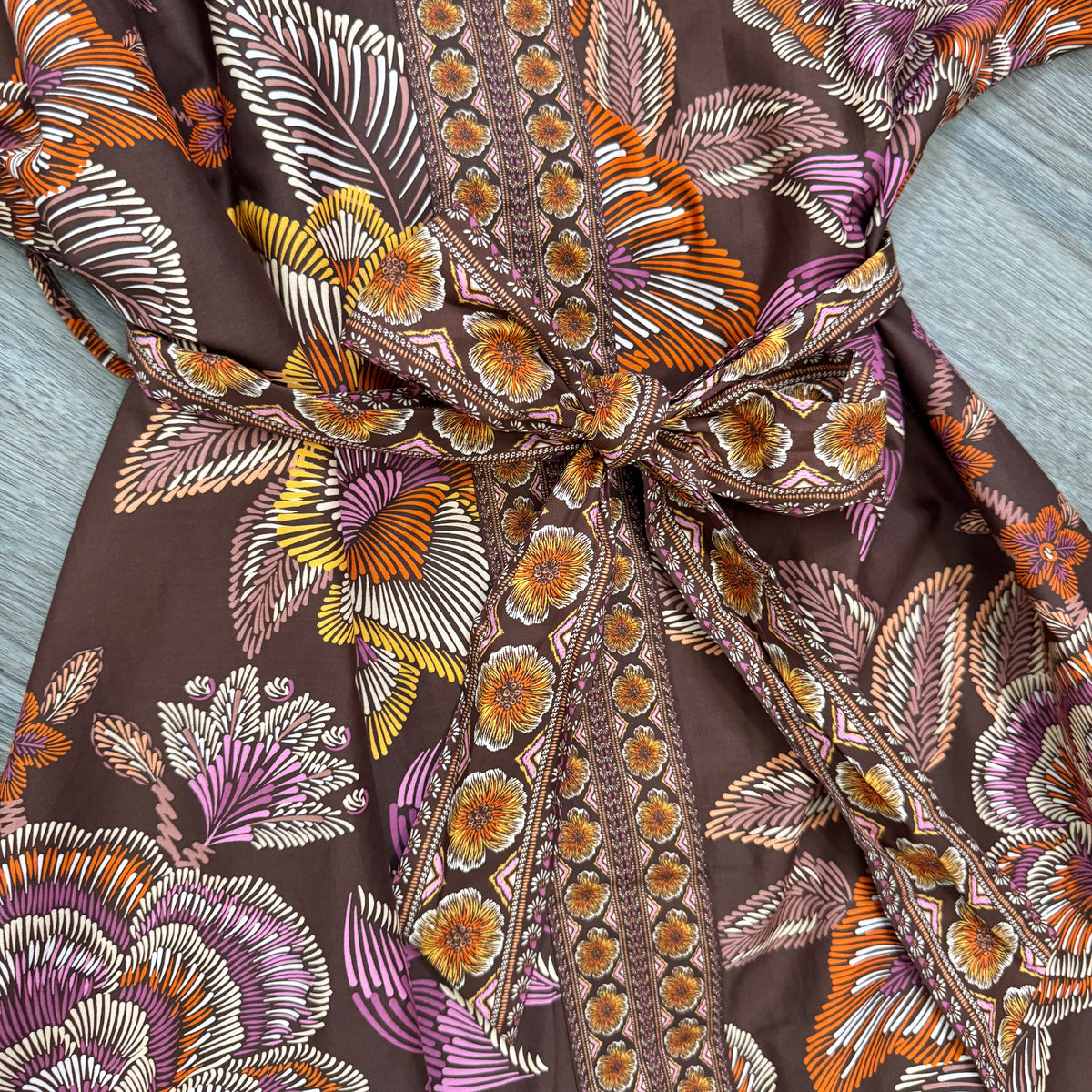 A close-up of a brown fabric garment with a bold botanical print in orange, yellow, and brown featuring a tie-waist detail.