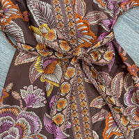 A close-up of a brown fabric garment with a bold botanical print in orange, yellow, and brown featuring a tie-waist detail.
