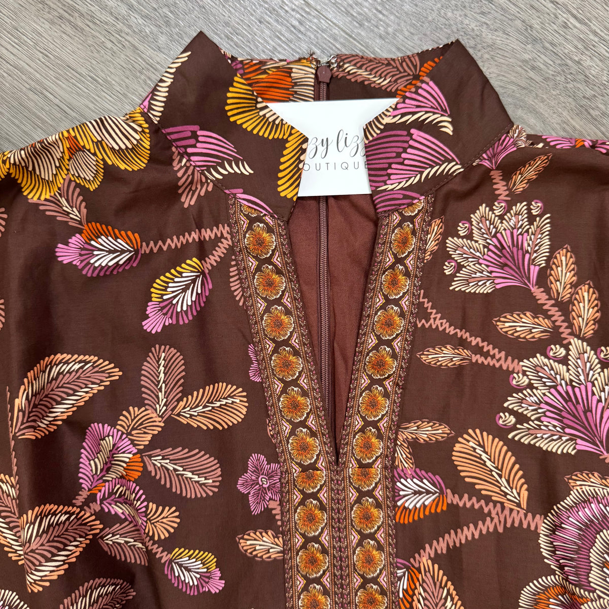 A close-up of a brown bohemian-style blouse with a colorful floral and leaf pattern in pink, orange, yellow, and beige, featuring a V-neckline with embroidered detailing and a white neck label reading bizzy lizzy boutique.