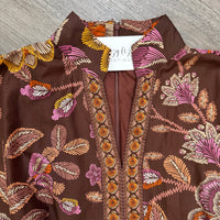 A close-up of a brown bohemian-style blouse with a colorful floral and leaf pattern in pink, orange, yellow, and beige, featuring a V-neckline with embroidered detailing and a white neck label reading bizzy lizzy boutique.