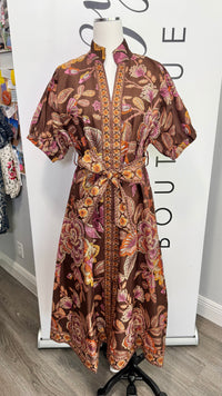 A brown short-sleeve kaftan dress with a vibrant floral and paisley print in pink, orange, and brown, displayed on a mannequin in front of a boutique backdrop.