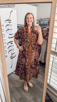 A woman takes a mirror selfie in a boutique wearing a brown floral maxi dress with a tie waist and short sleeves. The boutique interior is visible, including clothing racks and a white sign that reads 'Boutique'.