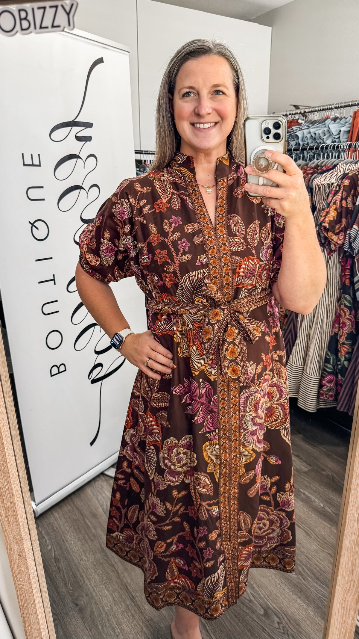 A woman takes a mirror selfie wearing a brown short-sleeve button-up dress with a vibrant floral and paisley pattern in orange, pink, and gold. The background shows a boutique interior with a white sign reading 'Boutique' and clothing racks.