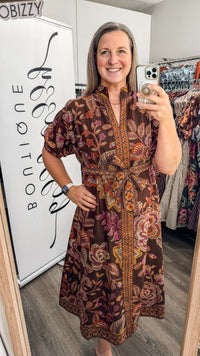 A woman takes a mirror selfie wearing a brown short-sleeve button-up dress with a vibrant floral and paisley pattern in orange, pink, and gold. The background shows a boutique interior with a white sign reading 'Boutique' and clothing racks.