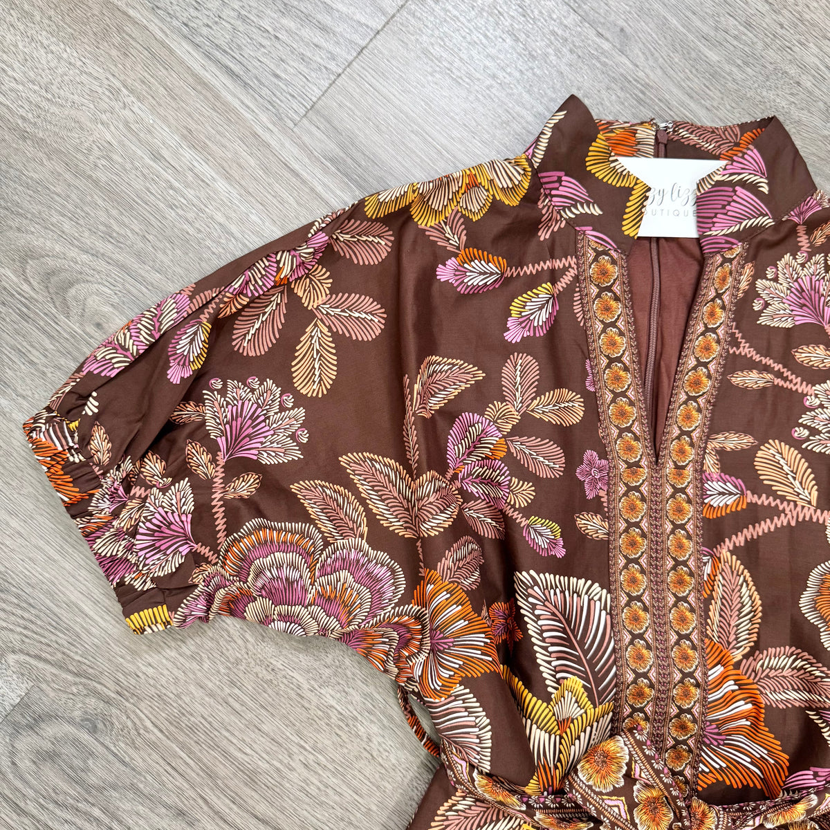 A brown short-sleeve blouse with a colorful floral and leaf print in pink, orange, and yellow, laid flat on a gray wood-look floor. A white neck label is visible.
