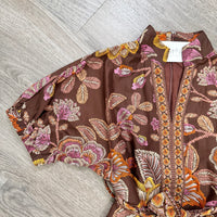 A brown short-sleeve blouse with a colorful floral and leaf print in pink, orange, and yellow, laid flat on a gray wood-look floor. A white neck label is visible.