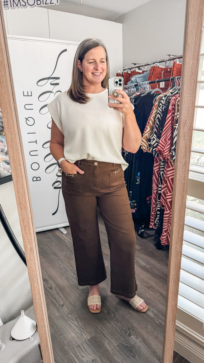 A woman takes a mirror selfie in a boutique showing off a cream-colored sleeveless top and brown wide-leg pants. The boutique interior is visible in the background with clothing racks and a wooden floor.