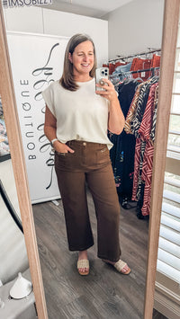 A woman takes a mirror selfie in a boutique showing off a cream-colored sleeveless top and brown wide-leg pants. The boutique interior is visible in the background with clothing racks and a wooden floor.