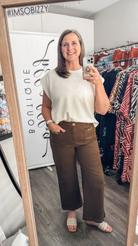 A woman takes a mirror selfie in a boutique showing off a cream-colored sleeveless top and brown wide-leg pants. The boutique interior features clothing racks with various garments and a white wall in the background.