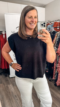 A woman takes a mirror selfie in a clothing store, wearing a black short-sleeve top and white pants.