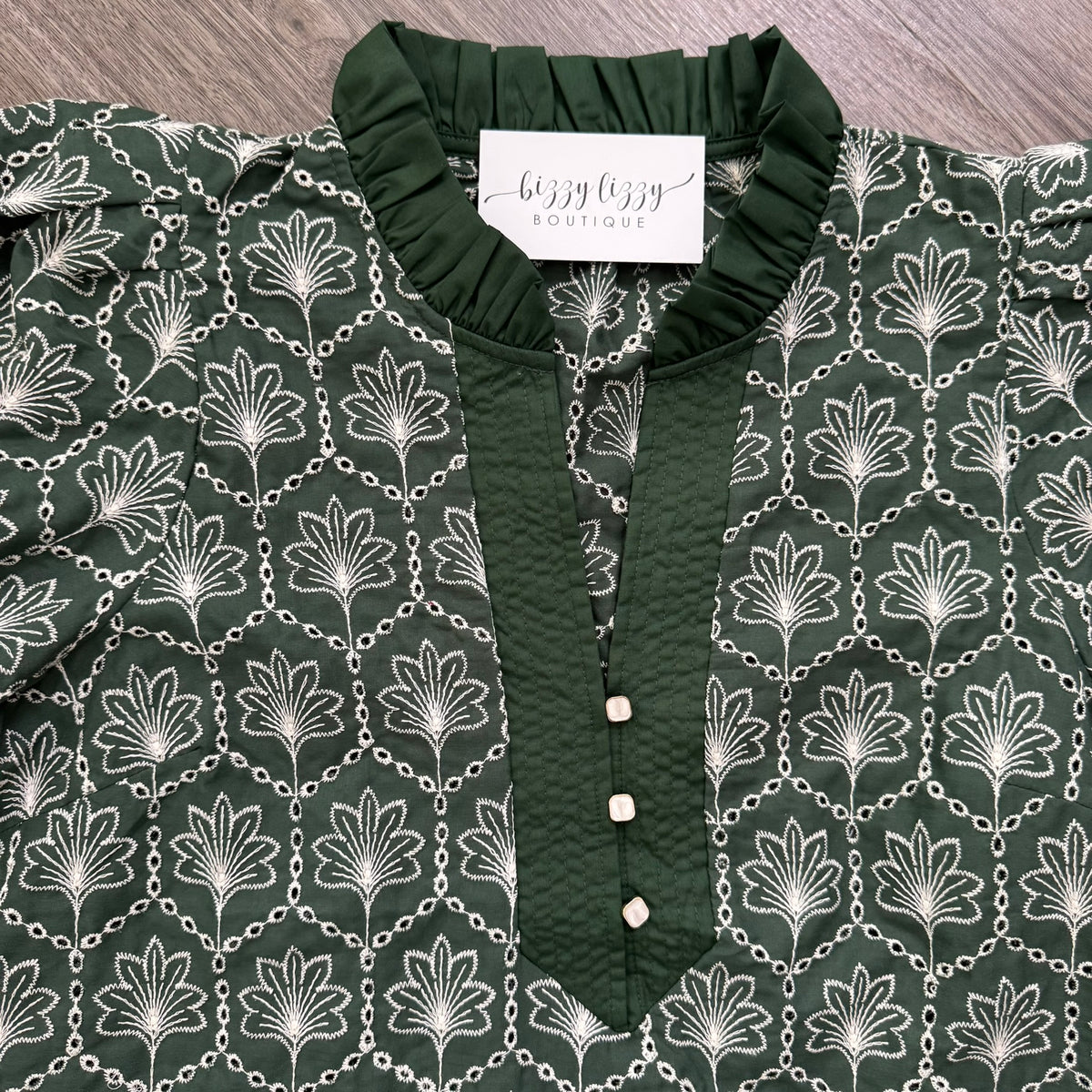 A dark green blouse with a white palm tree and chain pattern, featuring a solid green ruffled collar and button placket. A white label reading 'buzzy boutique' is visible inside the collar, and the garment is laid flat on a gray wood-textured surface.
