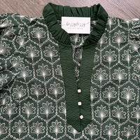 A dark green blouse with a white palm tree and chain pattern, featuring a solid green ruffled collar and button placket. A white label reading 'buzzy boutique' is visible inside the collar, and the garment is laid flat on a gray wood-textured surface.