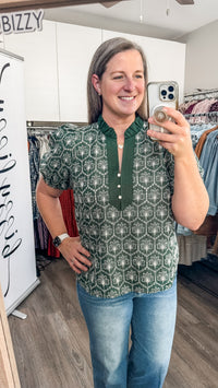 A woman takes a mirror selfie in a boutique while wearing a short-sleeve button-front top with a green and white geometric embroidered pattern and blue jeans.