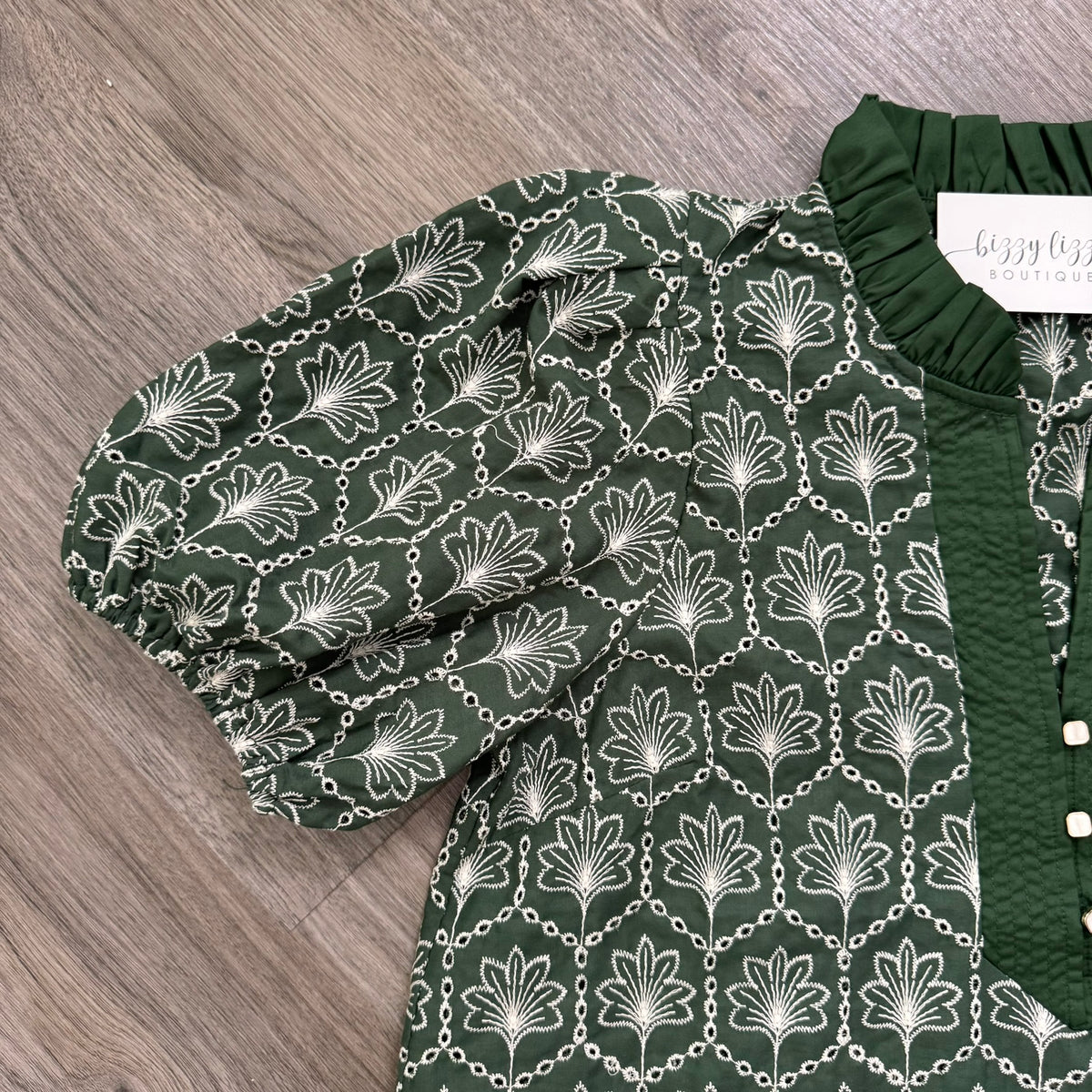 A dark green blouse with a white floral and chain pattern, featuring puff sleeves and a matching dark green ruffled collar, laid flat on a gray wood floor.