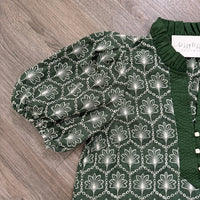 A dark green blouse with a white floral and chain pattern, featuring puff sleeves and a matching dark green ruffled collar, laid flat on a gray wood floor.
