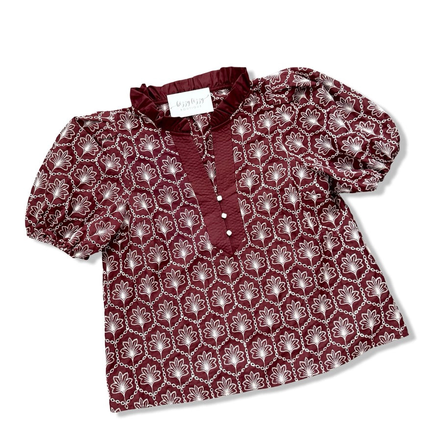 A women's short-sleeve blouse by Mignon, featuring a deep burgundy base with a repeating white floral pattern and a solid burgundy ruffled collar and button placket.