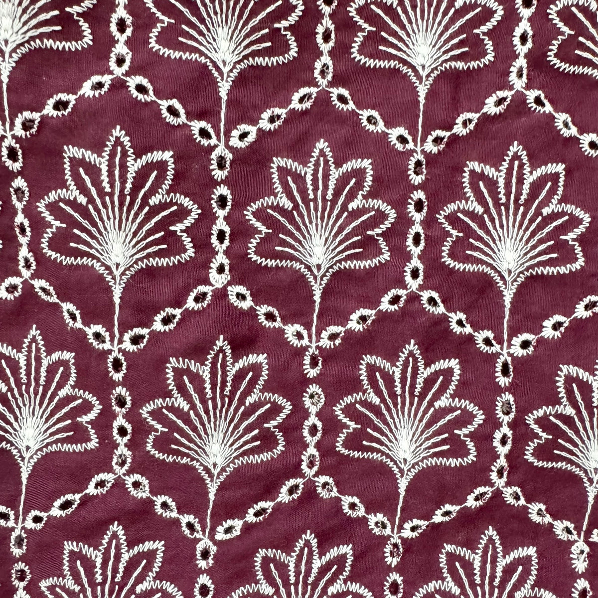 A close-up of a burgundy fabric with an intricate white embroidered floral and geometric pattern.