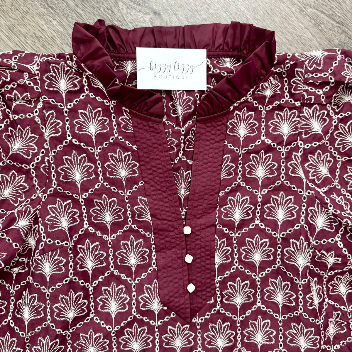 A close-up of a burgundy and white patterned blouse with a ruffled neckline, buttoned front, and repeating floral chain print, laid flat on a gray wood surface. A neck label reads 'bizzy lizzy boutique' with smaller text below.