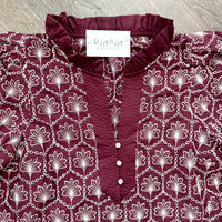 A close-up of a burgundy and white patterned blouse with a ruffled neckline, buttoned front, and repeating floral chain print, laid flat on a gray wood surface. A neck label reads 'bizzy lizzy boutique' with smaller text below.