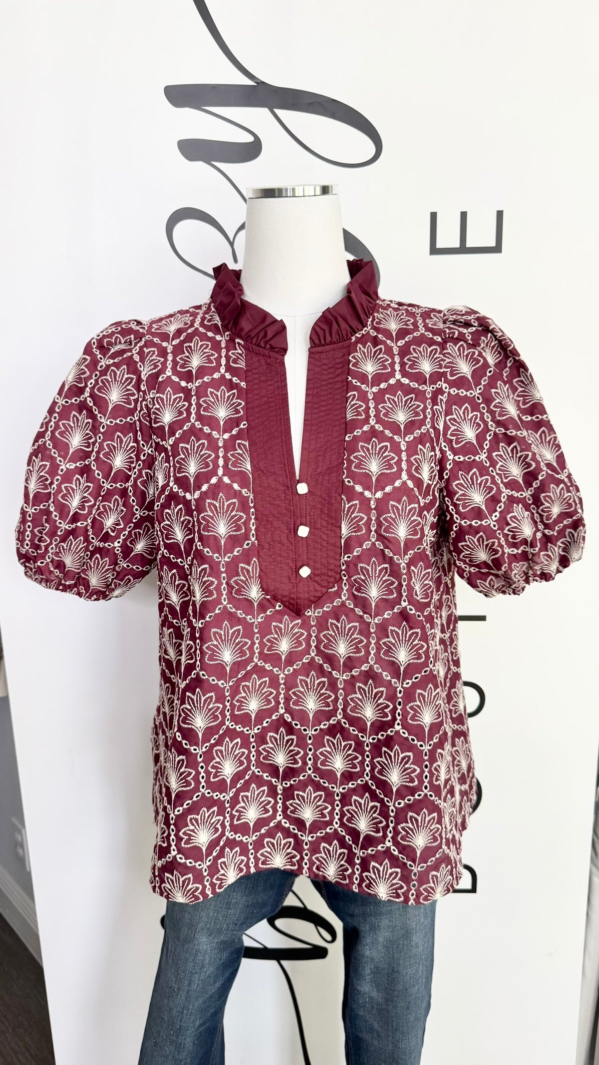 A short-sleeve blouse with puff sleeves and a ruffled high collar, featuring a burgundy and white geometric pattern, displayed on a mannequin torso with blue jeans against a white wall.