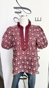 A short-sleeve blouse with puff sleeves and a ruffled high collar, featuring a burgundy and white geometric pattern, displayed on a mannequin torso with blue jeans against a white wall.