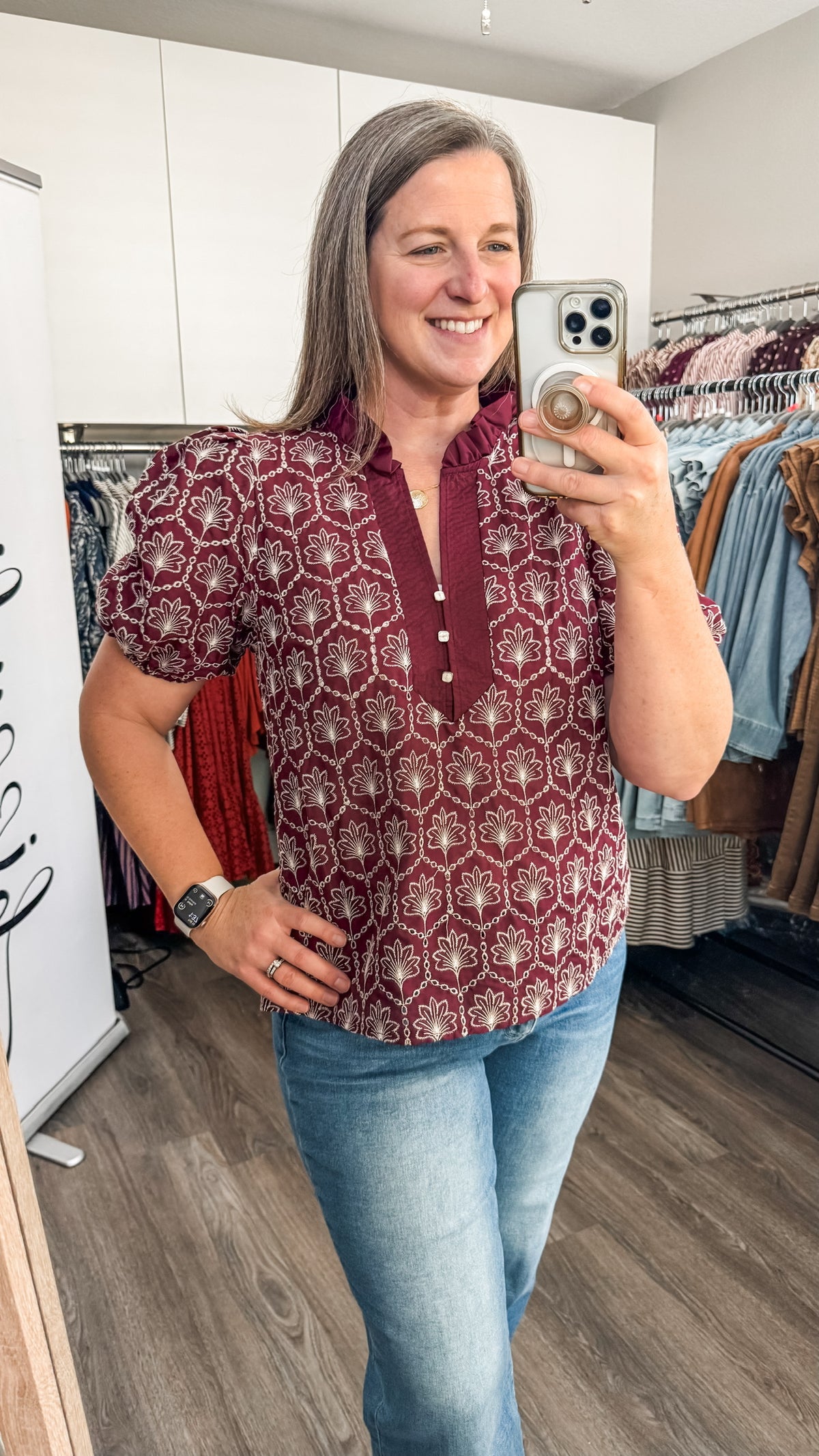 A woman takes a mirror selfie in a boutique while wearing a short-sleeve button-up blouse with a geometric embroidered pattern in burgundy and white, paired with blue jeans.
