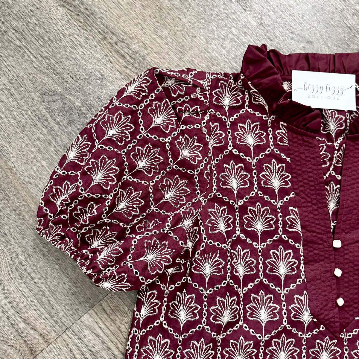 A close-up of a burgundy blouse with a white floral and chain pattern, featuring puff sleeves and a ruffled neckline, laid flat on a gray wood floor. A white brand tag reading 'bizzy lizzy boutique' is visible.