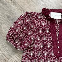 A close-up of a burgundy blouse with a white floral and chain pattern, featuring puff sleeves and a ruffled neckline, laid flat on a gray wood floor. A white brand tag reading 'bizzy lizzy boutique' is visible.