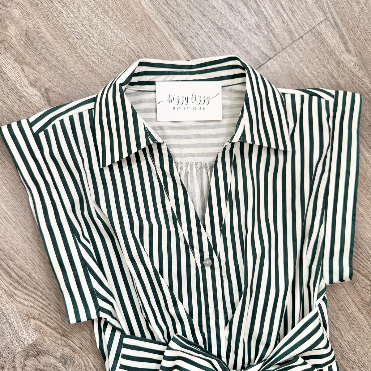A short-sleeve collared button-front shirt with vertical dark green and off-white stripes, laid flat on a light wood floor. The neck label reads 'bizzy lizzy boutique'.