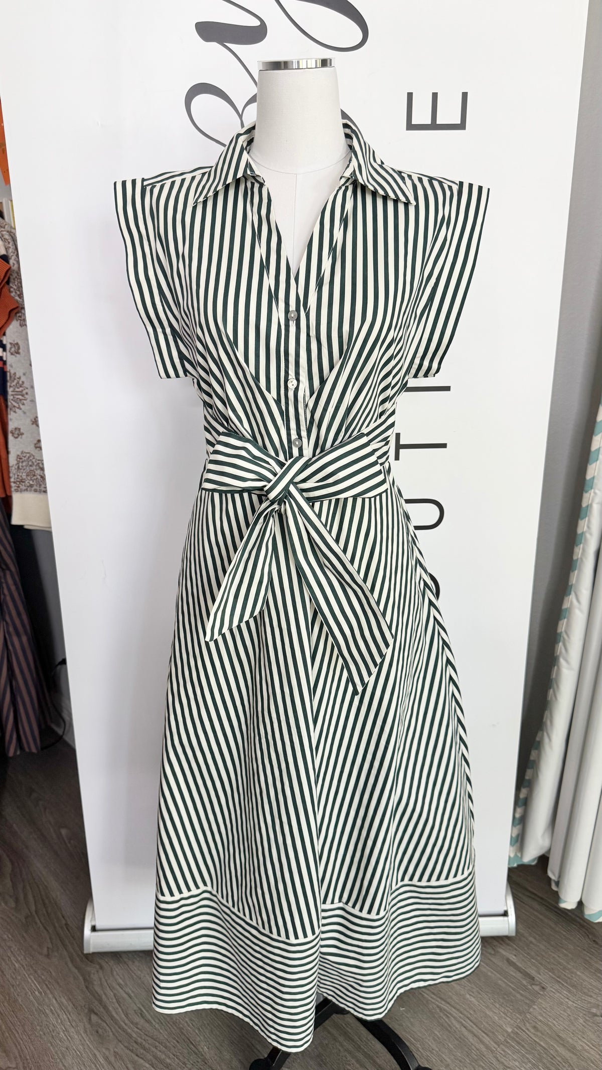 A sleeveless collared dress with a black and white striped pattern, featuring a self-tie waist belt, displayed on a mannequin in front of a white boutique backdrop.