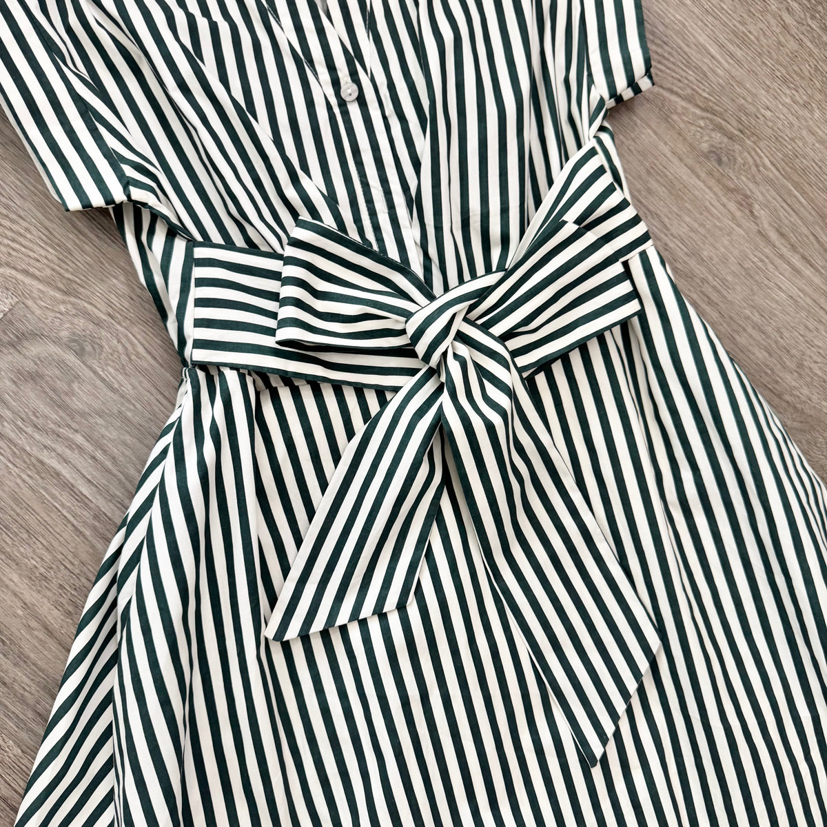 A close-up of a short-sleeve button-front dress with vertical dark green and off-white stripes, featuring a matching fabric tie belt at the waist, laid flat on a wood floor.