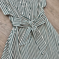 A close-up of a short-sleeve button-front dress with vertical dark green and off-white stripes, featuring a matching fabric tie belt at the waist, laid flat on a wood floor.