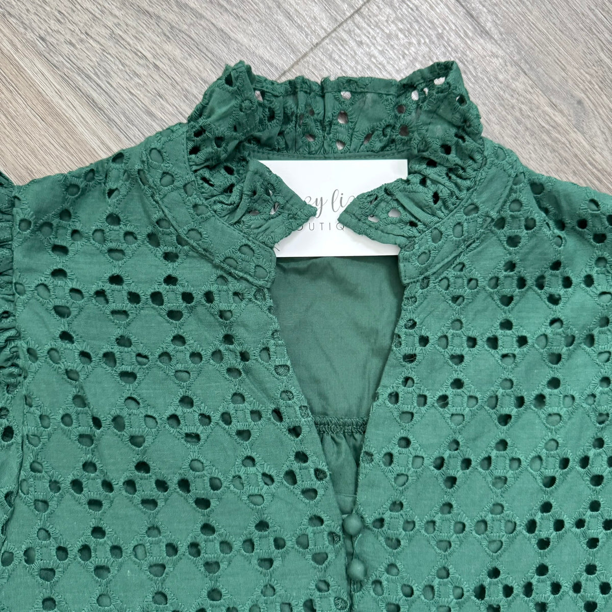 A green eyelet lace blouse with a ruffled mock neck and button-front closure, laid flat on a gray wood-look floor. A white brand tag is visible inside the collar.