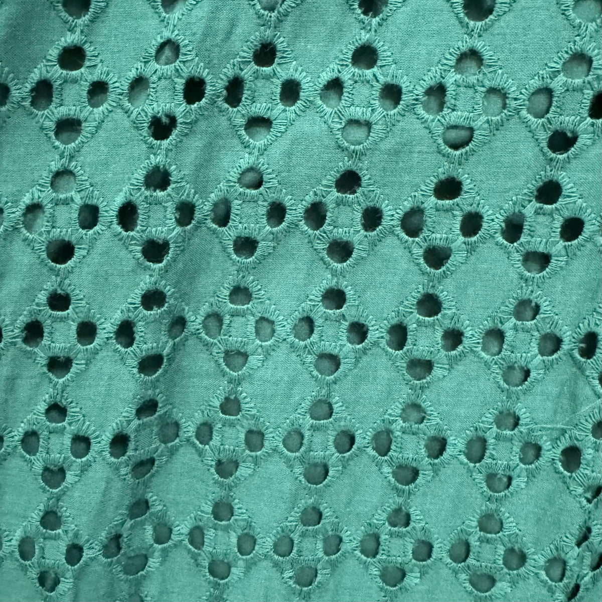 A close-up of a teal green eyelet fabric with a repeating diamond pattern of embroidered borders and circular cutouts.