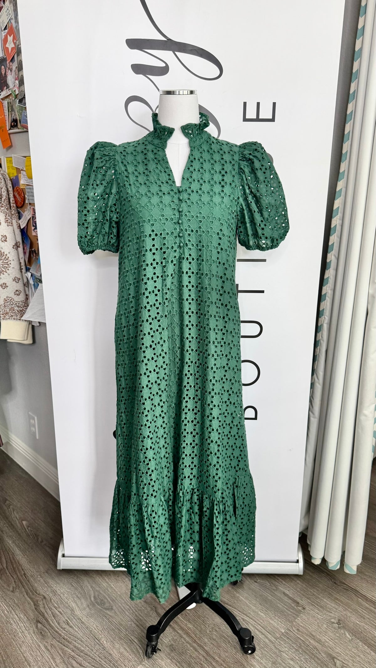 A green eyelet midi dress with puff sleeves and a high mock neck, displayed on a mannequin in front of a boutique backdrop.