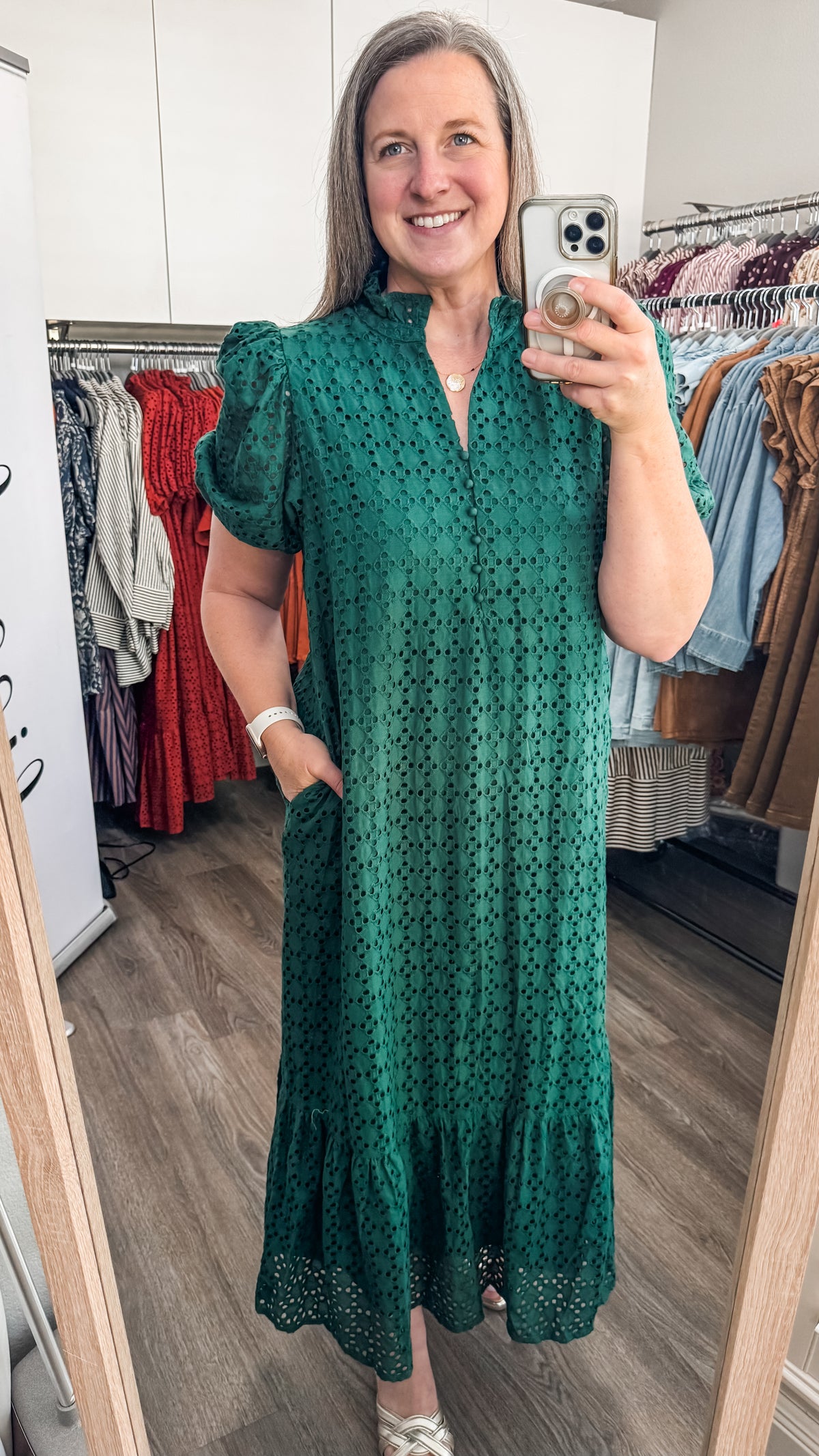 A woman takes a mirror selfie in a boutique while wearing a dark green eyelet midi dress with puff sleeves and a ruffled hem. Clothing racks are visible in the background.