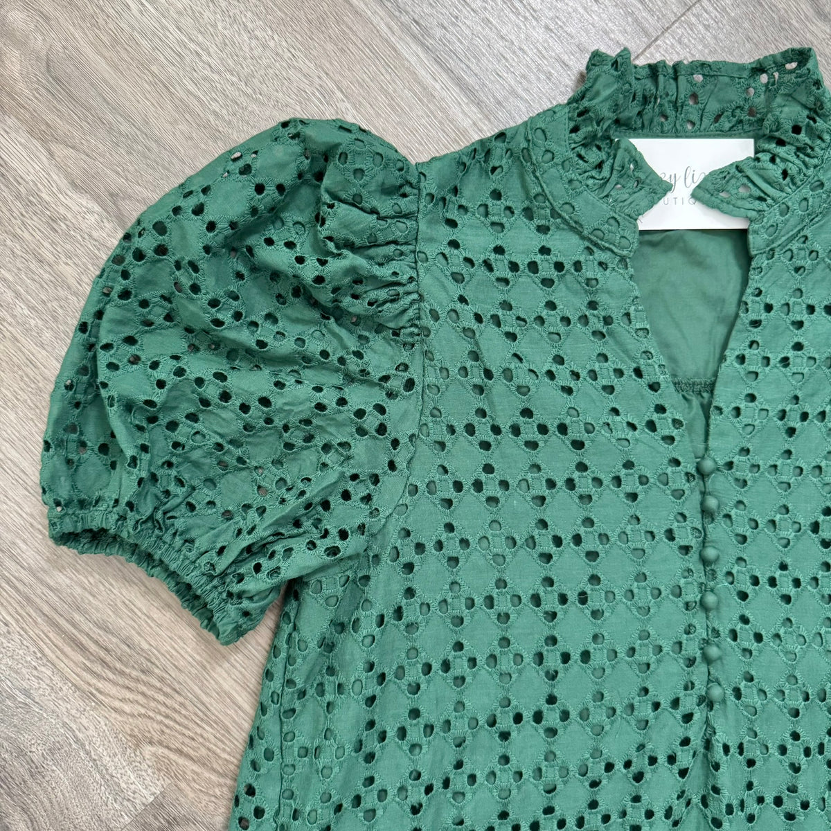 A green eyelet blouse with puff sleeves and a ruffled high neckline, laid flat on a light wood floor. The blouse features a button-down front and an allover eyelet pattern.