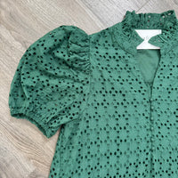 A green eyelet blouse with puff sleeves and a ruffled high neckline, laid flat on a light wood floor. The blouse features a button-down front and an allover eyelet pattern.