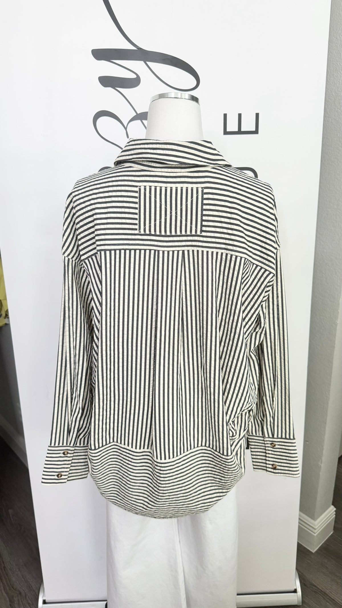 A black and white vertically striped button-down shirt with a high collar, displayed on a white mannequin torso in front of a white branded backdrop.