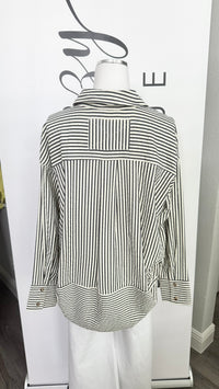 A black and white vertically striped button-down shirt with a high collar, displayed on a white mannequin torso in front of a white branded backdrop.