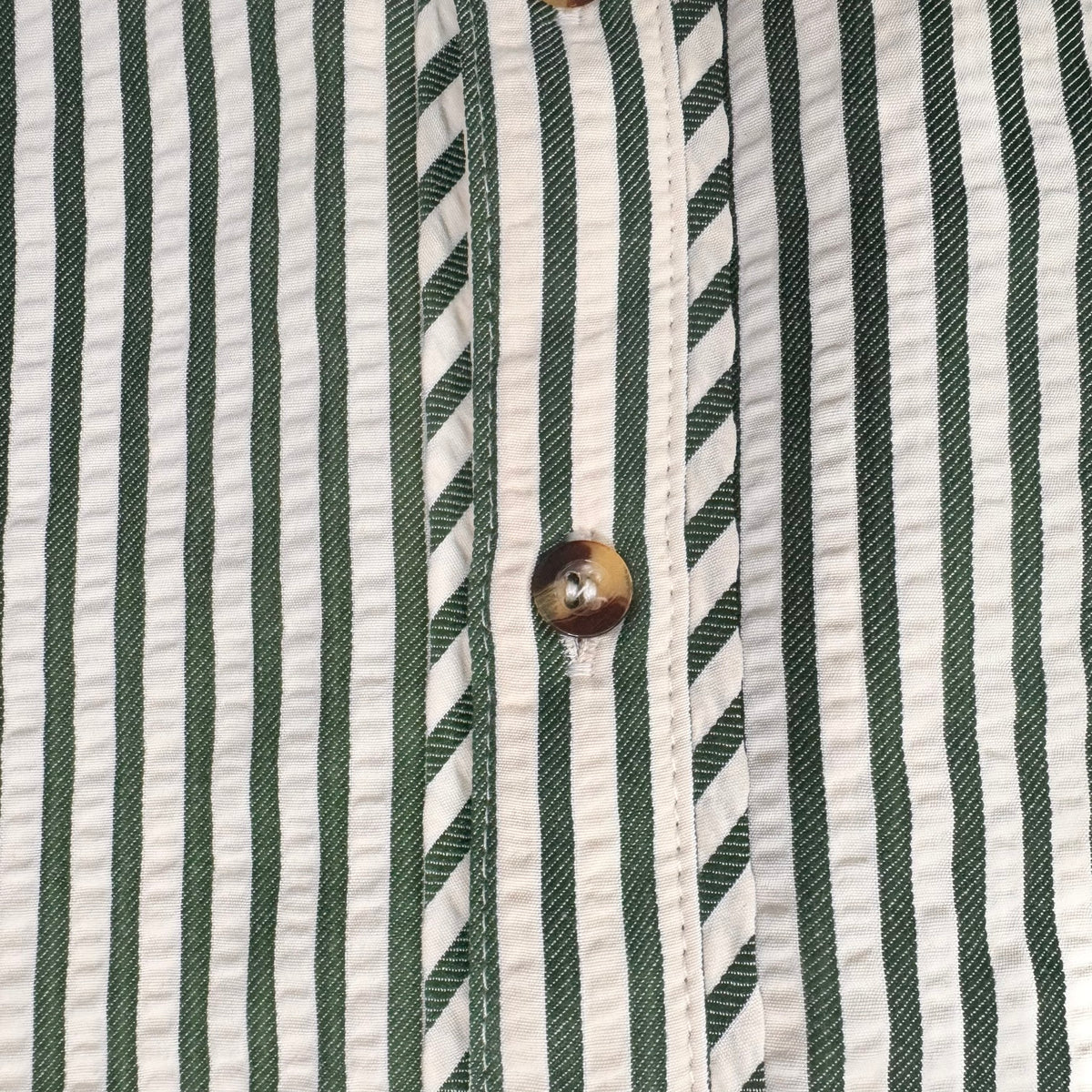 Close-up of a striped shirt fabric with vertical green and white stripes, featuring a brown button on the front placket.