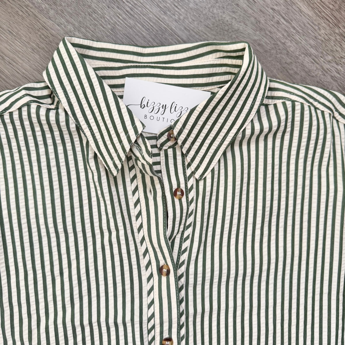 A close-up of a button-down shirt with vertical green and off-white stripes, featuring a white neck label that reads 'bizzy boutique' and brown buttons.
