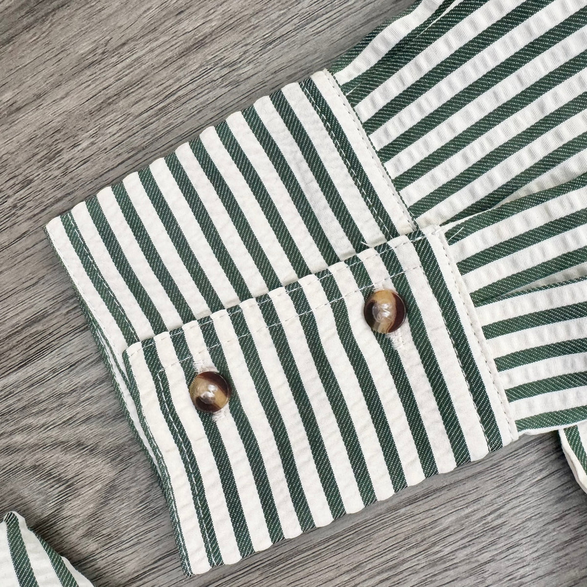 Close-up of a striped fabric garment laid flat on a gray wood surface, showing green and off-white vertical stripes with two brown shell-like buttons on the cuff.