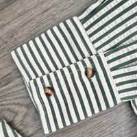 Close-up of a striped fabric garment laid flat on a gray wood surface, showing green and off-white vertical stripes with two brown shell-like buttons on the cuff.