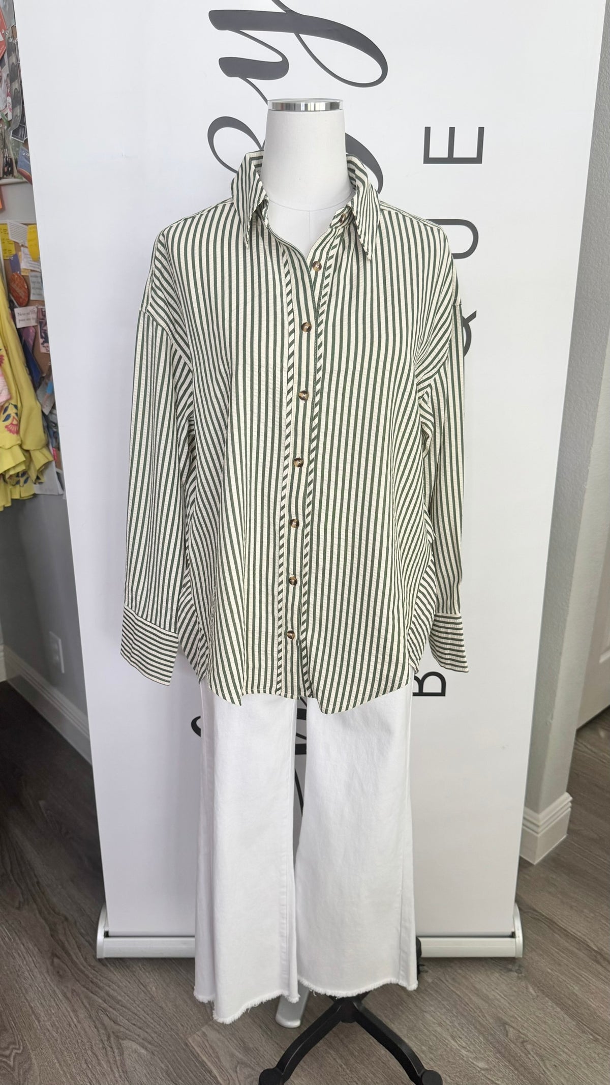 A long-sleeve button-up shirt with vertical green and white stripes, styled on a white mannequin torso with white wide-leg pants in front of a white boutique backdrop.