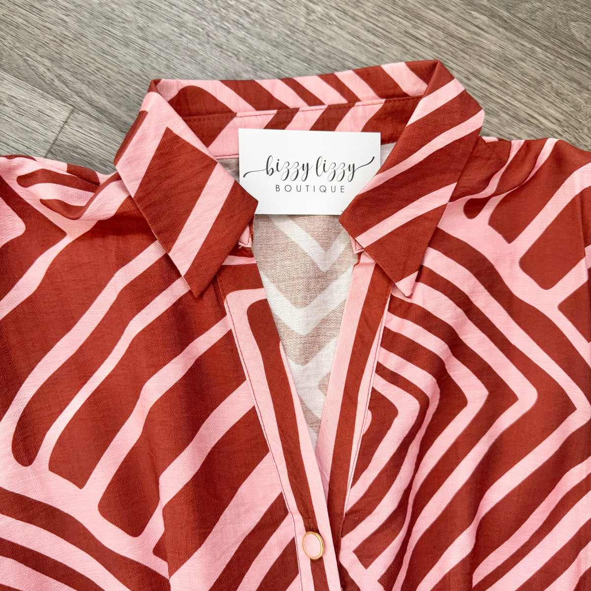 A close-up of a collared button-front blouse laid flat on a gray wood surface. The fabric has a bold geometric stripe pattern in terracotta red and light pink, with a visible white neck label reading 'bizzy lizzy BOUTIQUE'.