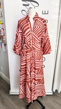 A long-sleeve collared button-front dress with a geometric wavy stripe pattern in terracotta red and white, displayed on a mannequin torso in an indoor boutique setting.